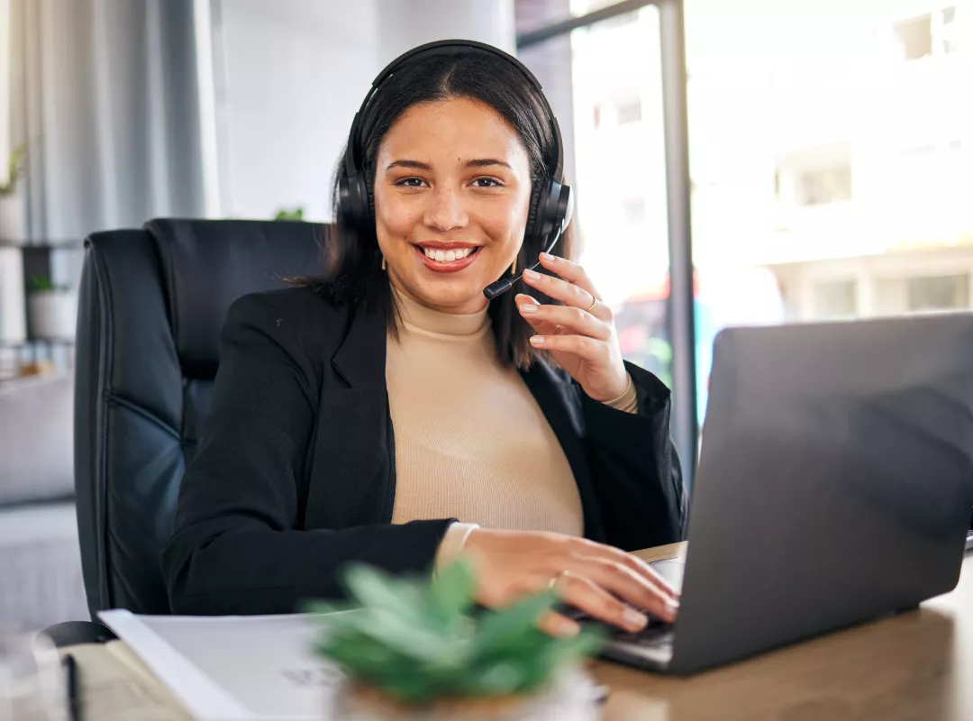 Woman wearing a headset smiles while sitting at a desk and using a laptop, appearing to engage in a video call or virtual meeting—ideal for appointment booking at aesthetic practices.